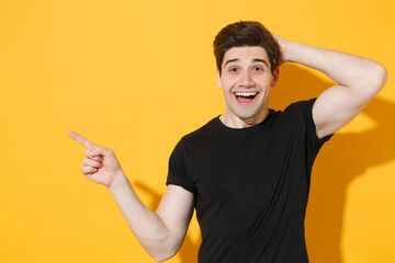 Excited young man guy in casual black t-shirt posing isolated on yellow wall background studio portrait. People lifestyle concept. Mock up copy space. Pointing index finger aside up, put hand on head.