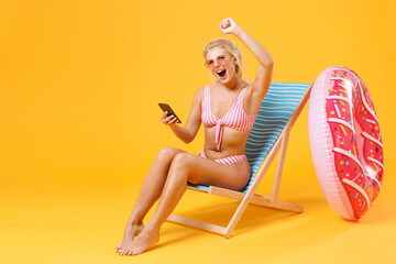 Screaming young woman girl in pink striped swimsuit glasses sit on deck chair isolated on yellow background. People summer vacation rest lifestyle concept. Using mobile phone, doing winner gesture.