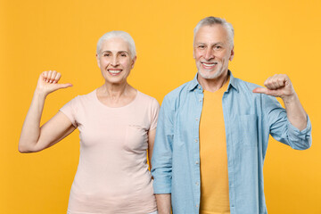 Smiling elderly gray-haired couple woman man in casual clothes posing isolated on yellow background studio portrait. People emotions lifestyle concept. Mock up copy space. Point thumbs on themselves.