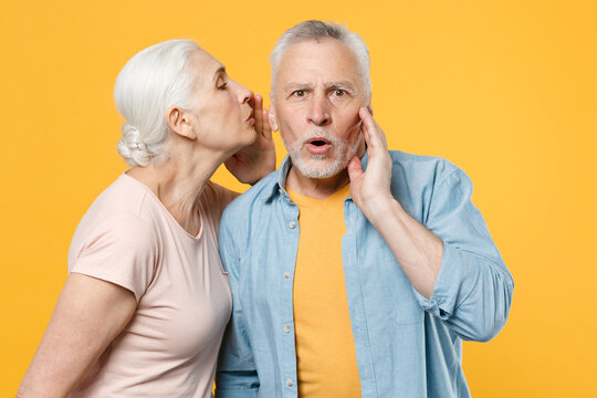 Shocked elderly gray-haired couple woman man in casual clothes isolated on yellow background studio portrait. People lifestyle concept. Mock up copy space. Whispering secret behind hand, sharing news.