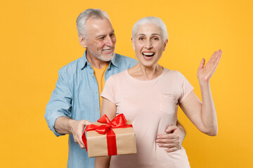 Excited elderly gray-haired couple woman man in casual clothes isolated on yellow background studio. St. Valentine's Day, Women's Day, birthday, holiday concept. Hold present box with gift ribbon bow.