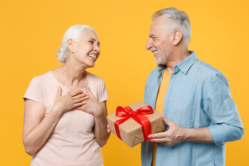Smiling elderly gray-haired couple woman man in casual clothes isolated on yellow background studio. St. Valentine's Day, Women's Day, birthday, holiday concept. Hold present box with gift ribbon bow.