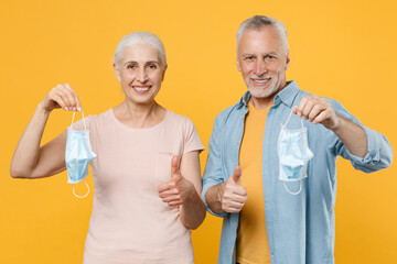 Smiling gray-haired couple woman man in casual clothes isolated on yellow background. Epidemic pandemic coronavirus 2019-ncov sars covid-19 flu virus concept. Hold sterile face mask showing thumbs up.