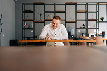 Laughing cheerful young man wearing fashion casual clothing is writing text on paper while sitting at the wooden desk in modern room. Concept of creative or office working.