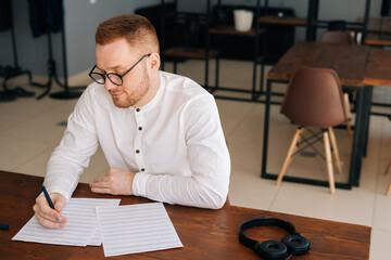 Talented young composer wearing stylish eyeglasses writing musical notes with pen on paper sheet music sitting at the wooden desk in modern room in office or home, headphones on table