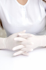 Hands in protective white medical gloves of the woman sitting at the white table. Crossed fingers in gloves