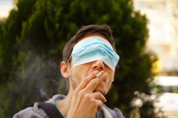 A portrait of a man with a face mask on his eyes. He is smoking a cigarette.