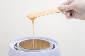 Wax heater with hot yellow wax on wooden spatula in human hand. Hot wax for depilation. Salon...