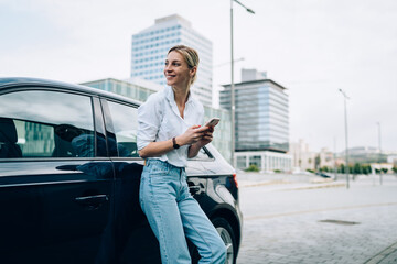 Optimistic woman with smartphone standing beside car and looking away