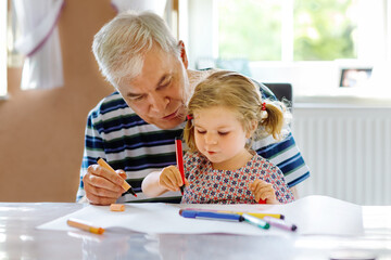 Cute little baby toddler girl and handsome senior grandfather painting with colorful pencils at...