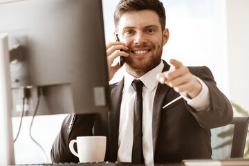 Business concept. Happy smiling young businessman sitting in office talking on a cell phone getting good news about his work and pointing with finger. Man in suit indoors on glass window background