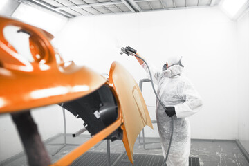 Man in protective mask and clothes sprays varnish to car hood with a spray gun in a paint booth.