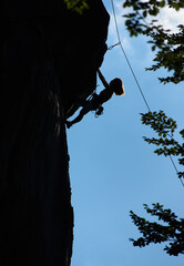 Silhouette of female alpinist climbing extremely vertical rock under blue sky. Young woman ascending high boulder and trying to reach mountaintop. Concept of mountaineering, alpinism, extreme sports.