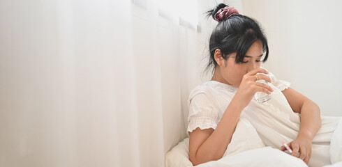 An ill woman drinks water and consuming a pill while lying on the bed in the bedroom.