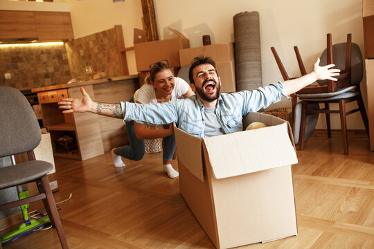 Young married couple moving into a new home.Man sitting in cardboard box while woman pushes him all over the room.Real estate funny concept
