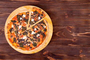 Pizza with cherry tomatoes, spinach, mozzarella, feta, kalamata olive and mushrooms, with a slice slightly removed on a wood platter which is on wooden rustic table, top view and copy space
