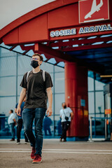 Fototapeta premium Young man dressed in black t-shirt, jeans, red sneakers, backpack, surgical mask and glasses walking away from the airport
