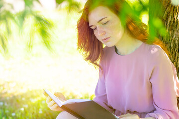 young, beautiful woman is reading a book in a park. Romantic red haired female outdoors.