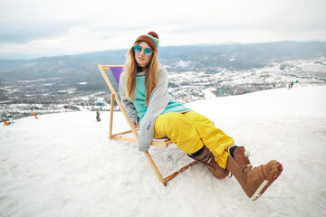 Snowboarder woman at winter mountains resting on sun-lounger at nice sun day. Sheregesh, Russia.