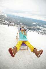 Snowboarder woman at winter mountains resting on sun-lounger at nice sun day. Sheregesh, Russia.