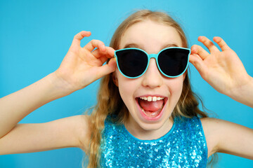 Beautiful mischievous Caucasian little girl in sunglasses close-up on a blue background. The girl is dressed in a beautiful blue dress. The concept of emotions and facial expressions.