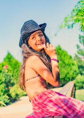 A pretty girl in a swimsuit and a black cowboy hat is relaxing on the beach by the sea. Smiling cute little girl on vacation. Charming girl looks to the side. Summer travel concept