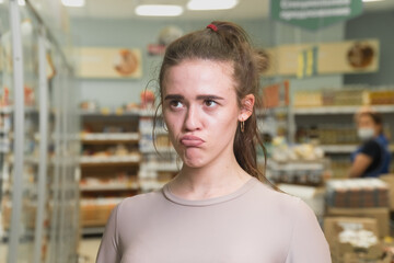 A sad upset girl in a super martette with a grocery cart. Portrait of a disgruntled buyer.