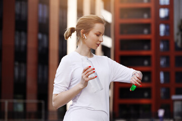 Woman in white t shirt standing on street  near building and skyscrapers in wireless headphones with smart watch. Drinking water out of bottle.  Doing sports, healthy lifestyle, training and workout.