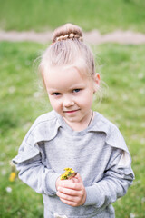 Little blond girl with blue eyes, hair arranged in a bun, wearing a gray dress, close-up portrait, looking into the camera, smiling, bored, playing on a lawn, sitting on the machine tires. Preschool  