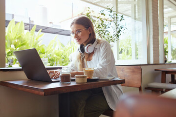 Woman. Online Work At Coffee Shop. Happy Girl In Casual Clothes With Laptop And Headphones Typing Something. Modern Digital Technologies For Remote Job Or Education At Comfortable Workplace.