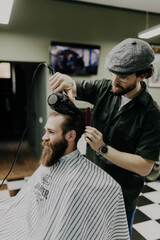 Close up side view of young bearded man getting groomed by hairdresser with hair dryer at barbershop