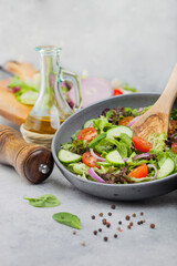 Fresh vegetarian vegetables salad with tomatoes and cucumber, lettuce and spinach in grey bowl with spatula on light board with olive oil, mill and fresh vegetables on round chopping board.