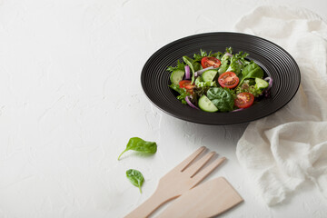 Fresh vegetarian vegetables salad with tomatoes and cucumber, lettuce and spinach in black bowl plate with spatula on light board with white kitchen towel.
