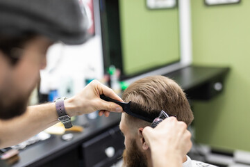 Young barber making haircut with razor to attractive bearded man in barbershop
