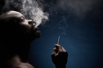 Fototapeta premium profile photo of a dark-skinned muscular handsome guy with a beard on a dark background who breathes out smoke from his mouth with a cigarette in his hand