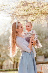 Smiling thirty-year-old woman holds beautiful baby girl in dress on a blurr background in a counter-light summer. mother and daughter