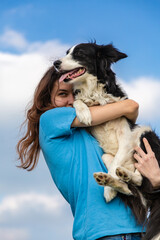 A girl with long hair in a blue T-shirt holds a black and white border collie dog in her arms. Portrait against a bright blue sky with clouds. Vertical orientation. 