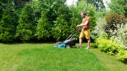 Mowing grass on the lawn with a lawn mower. A handsome adult man with an electric lawnmower with a grass catcher takes care of the lawn on a summer day. Summer lawn care