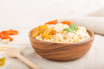 Bulgur porridge with dried apricots,  raisins and cashew in wooden bowl on a white wooden background and linen textile. Side view, selective focus.