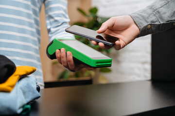 Client using smartphone and nfc terminal for contactless payment on counter in the clothes store. NFC and contactless technology concept. Modern shopping 