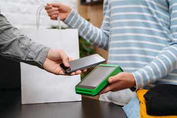 Contactless payment concept, male customer holding smartphone near nfc technology on counter, client make transaction, pay bill on terminal rfid cashier machine in clothes store. NFC technology
