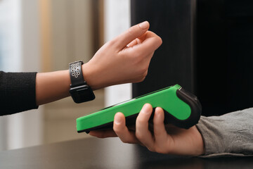 Closeup of modern lady paying via smartwatch while making purchase in shop. Woman customer using hand watch for contactless payment via payment terminal on counter in store. NFC technology concept