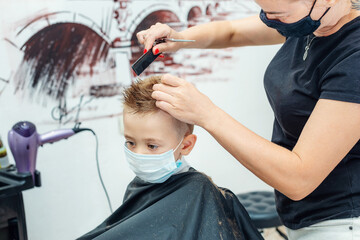 Caucasian little boy getting hair cut at the barbershop wearing protective mask. 