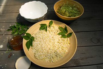 Harvest of shelled elderberry flowers, in a bamboo dish, on a grey wooden table. A jar of...