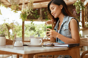 Business. Woman Drinks Smoothie At Cafe. Happy Asian Girl In Jeans Outfit With Laptop Enjoying Fresh Detox Cocktail. Modern Technologies For Digital Nomad Lifestyle And Online Work. 