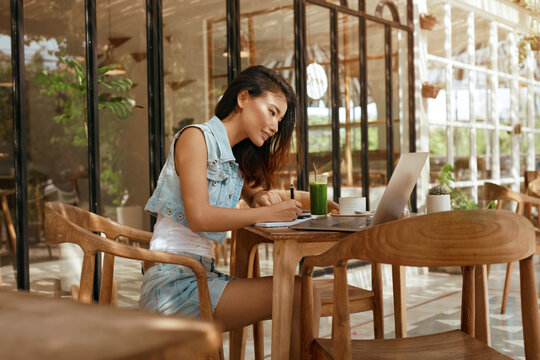 Work. Woman With Laptop At Coffee Shop. Asian Girl In Jeans Outfit Writing In Planner Book At Cafe. Remote Working Or Online Studying On Vacation With Modern Digital Technologies. 