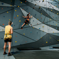 Male instructor holding rope, teaching, looking at woman in safety equipment and harness while she...