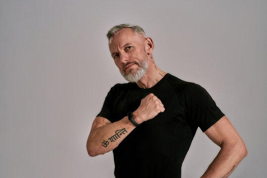 Middle aged muscular man in black t shirt looking at camera, beating his chest while posing in studio over grey background