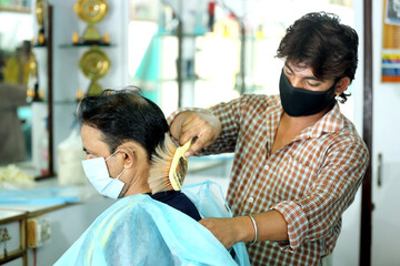 An Indian male hair dresser cutting hair in his beauty saloon with proper safety measures for...