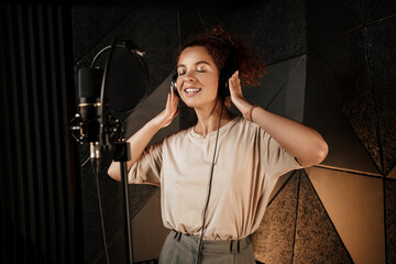 Beautiful smiling singer girl in headphones dreamily recording song in professional studio